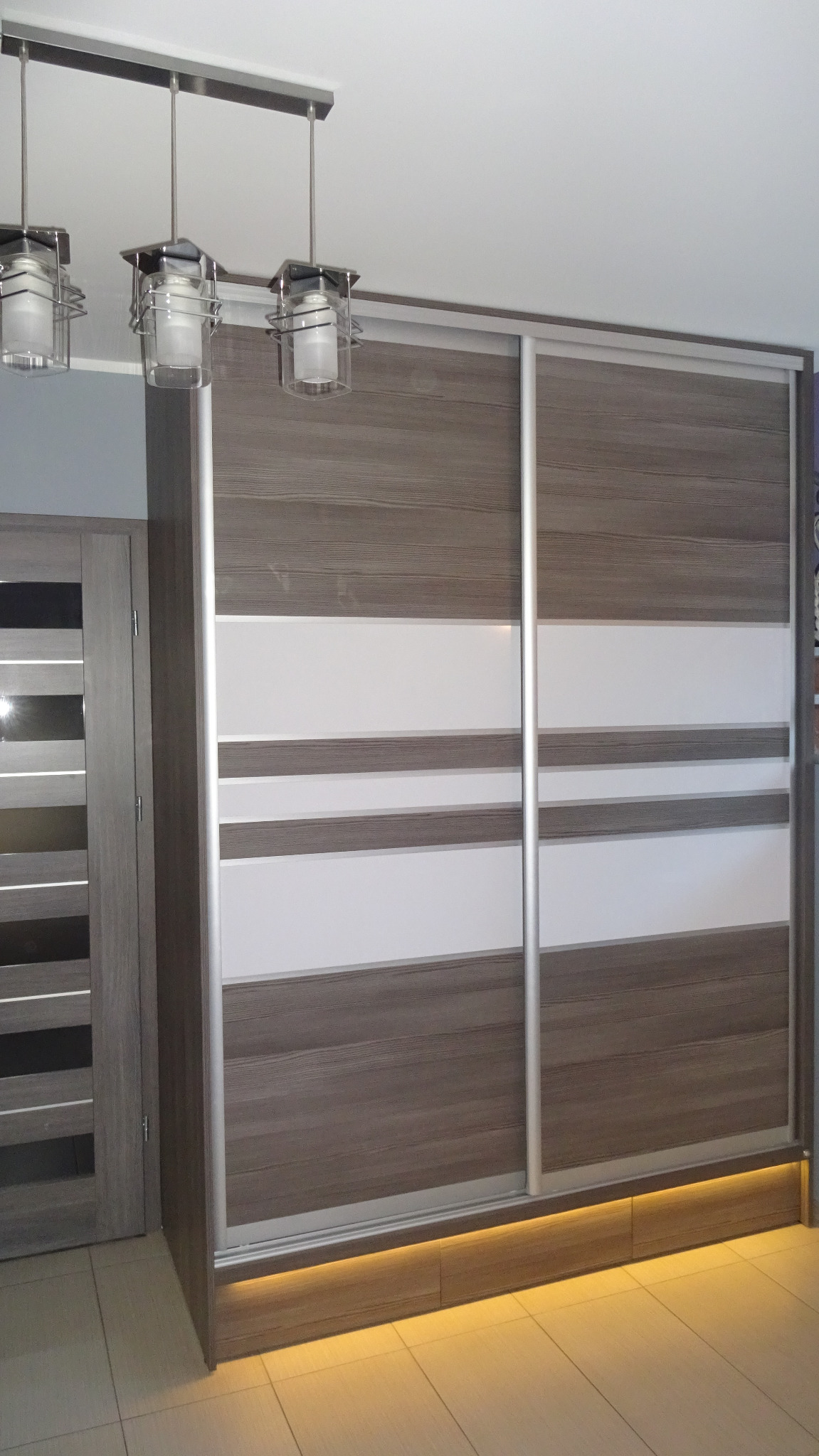 Szafa wnękowa z przesuwnymi drzwiami w odcieniach szarości i bieli, z subtelnym podświetleniem LED u dołu. Widoczne trzy lampy sufitowe.