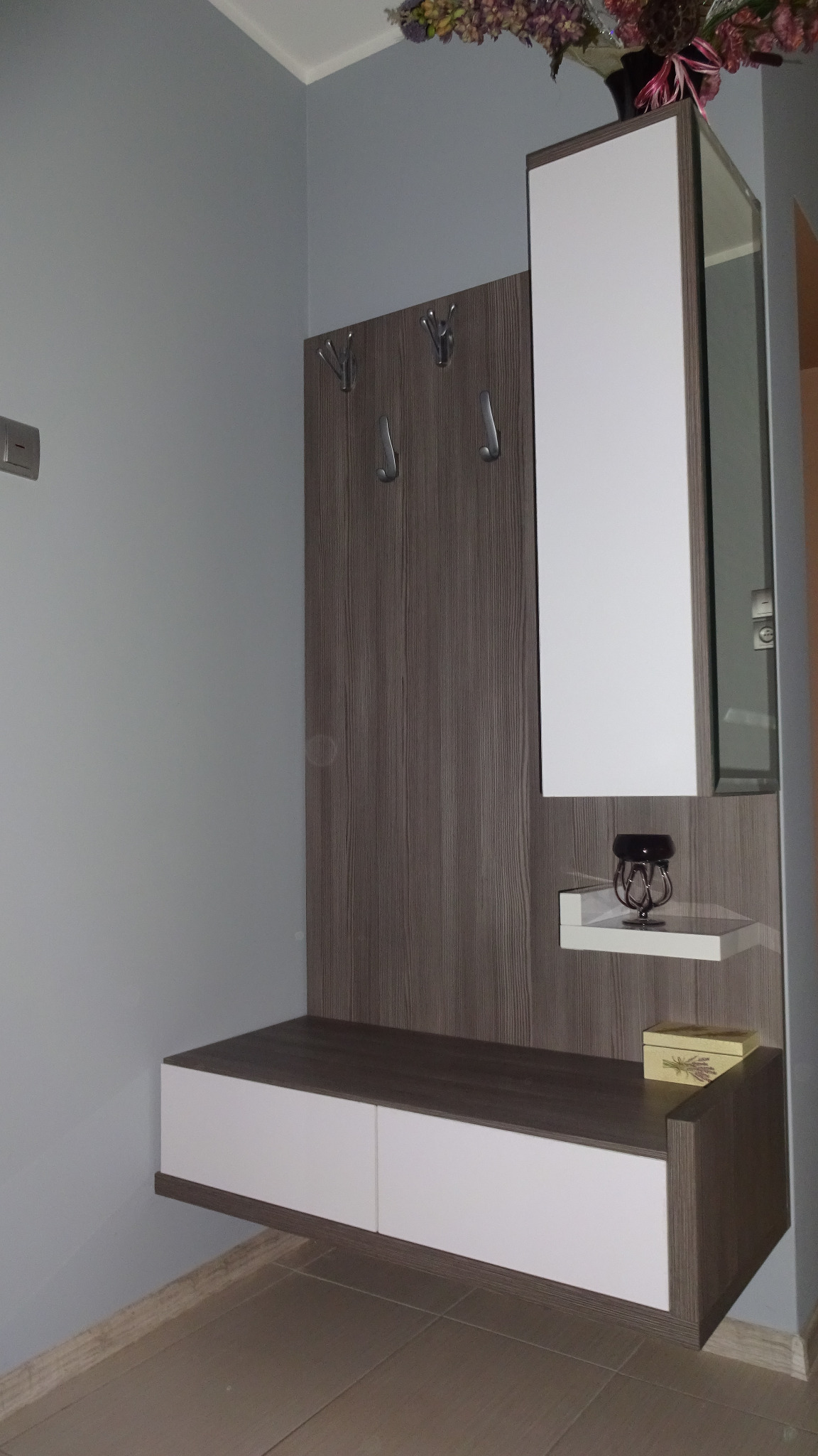 Nowoczesna szafa wnękowa z lustrem i wieszakami, wykończona w odcieniach bieli i drewna, uzupełniona o dekoracyjne akcenty, jak wazon z kwiatami i ozdobny świecznik.