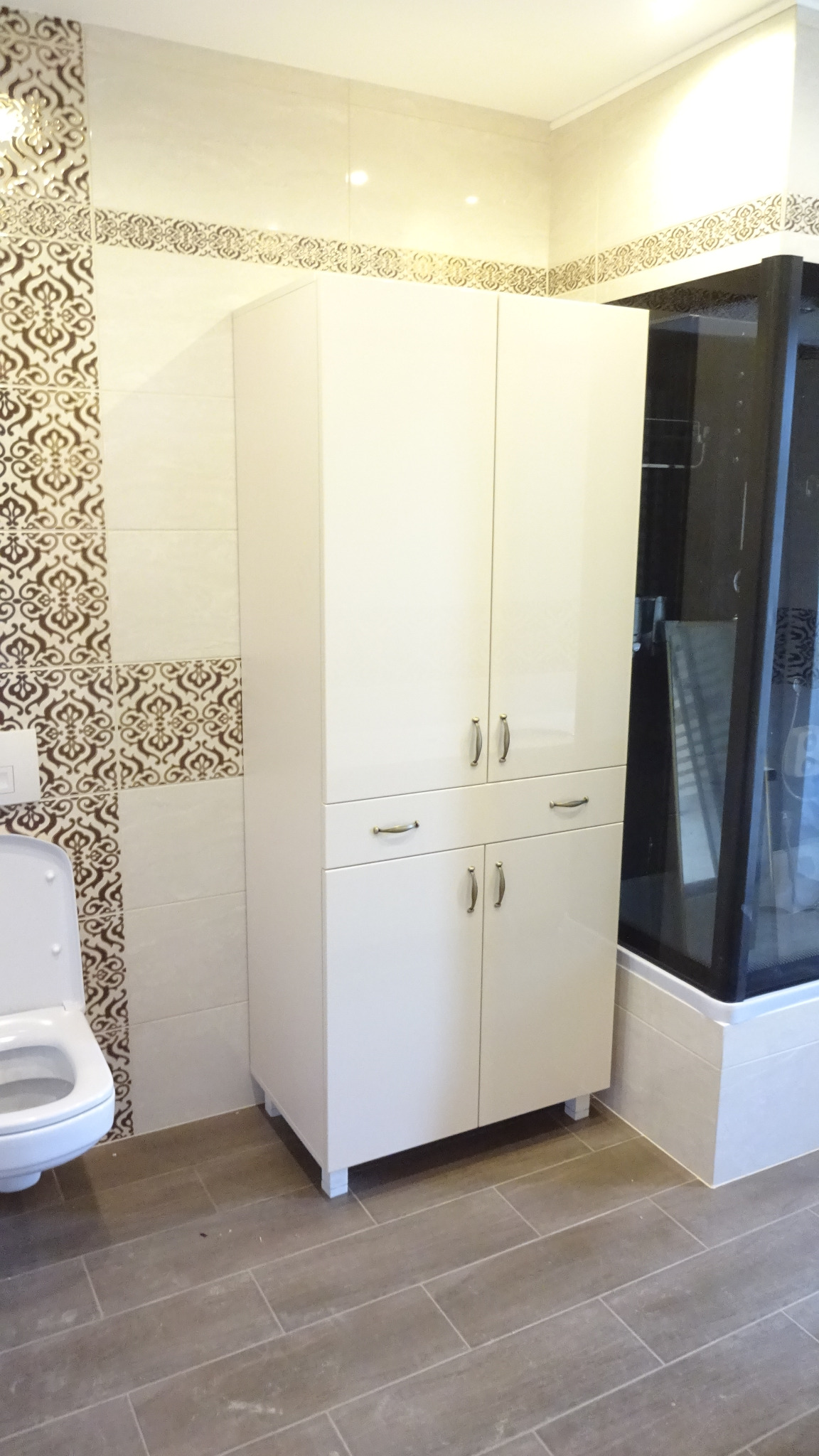 Biała szafka łazienkowa z czterema drzwiczkami i dwiema szufladami, z metalowymi uchwytami, na tle płytek z dekoracyjnym wzorem i kabiny prysznicowej.