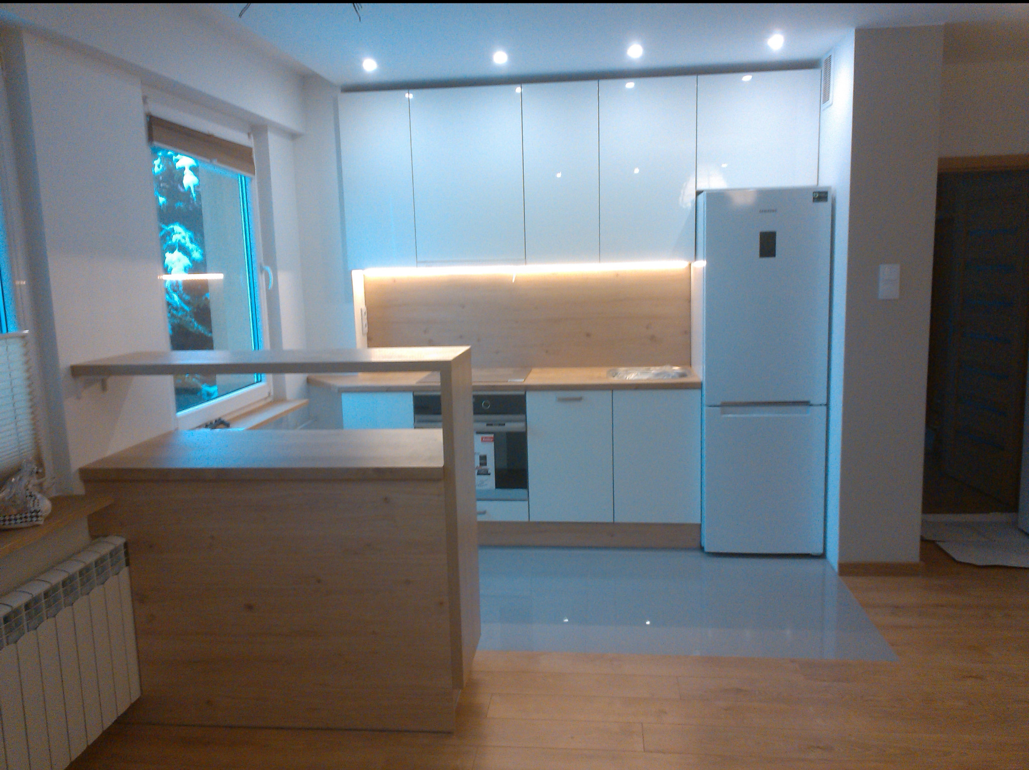 Nowoczesna kuchnia z białymi, błyszczącymi frontami szafek i drewnianym blatem. Widoczny barek z jasnego drewna i oświetlenie LED pod szafkami górnymi. Minimalistyczny design.