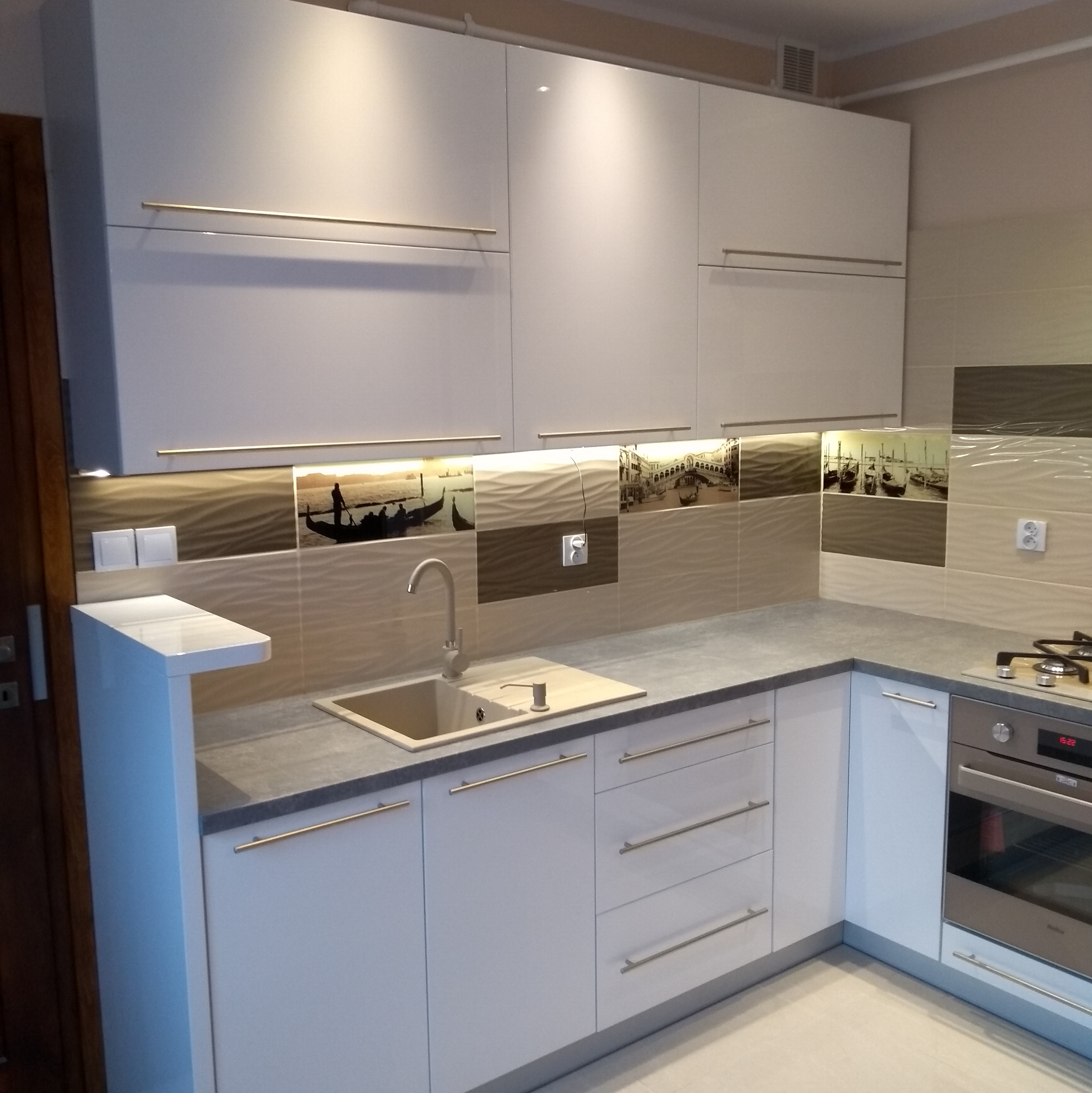 Biała zabudowa kuchenna z szarym blatem i złotymi uchwytami w Suchedniowie. Na ścianie dekoracyjne płytki z motywem weneckim. Nowoczesny design.