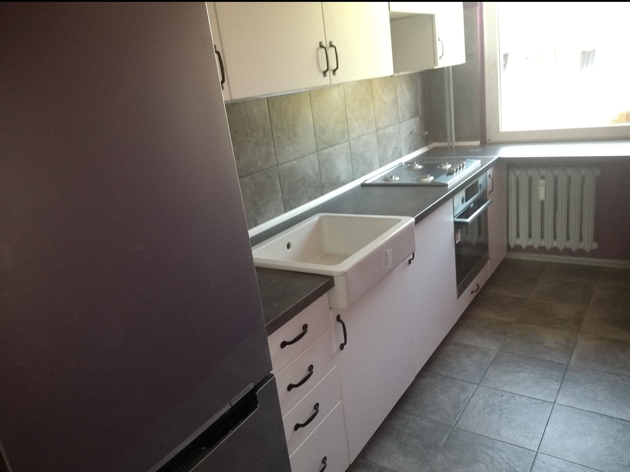 Biała kuchnia z ciemnym blatem i ceramicznym zlewem. Widoczny fragment lodówki, płyta grzewcza i piekarnik. Szare płytki na ścianie i podłodze. Ujęcie z niskiego kąta.