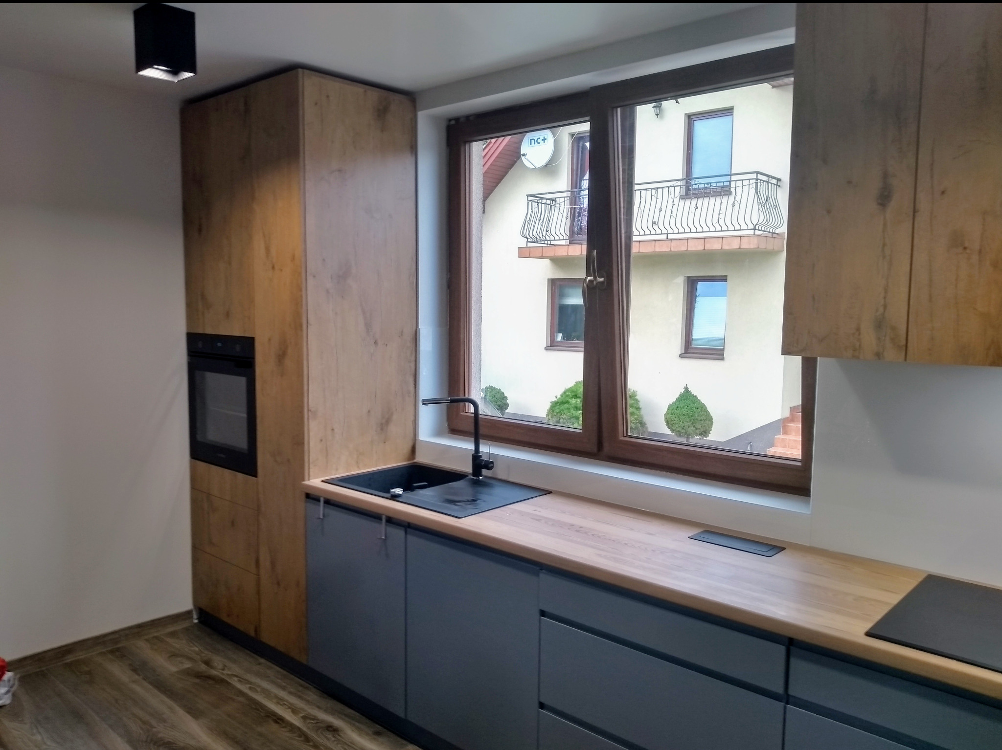 Nowoczesna kuchnia z szarymi szafkami i drewnianym blatem pod oknem, z widokiem na budynek z balkonem. Uwagę przykuwa czarny zlew i bateria.