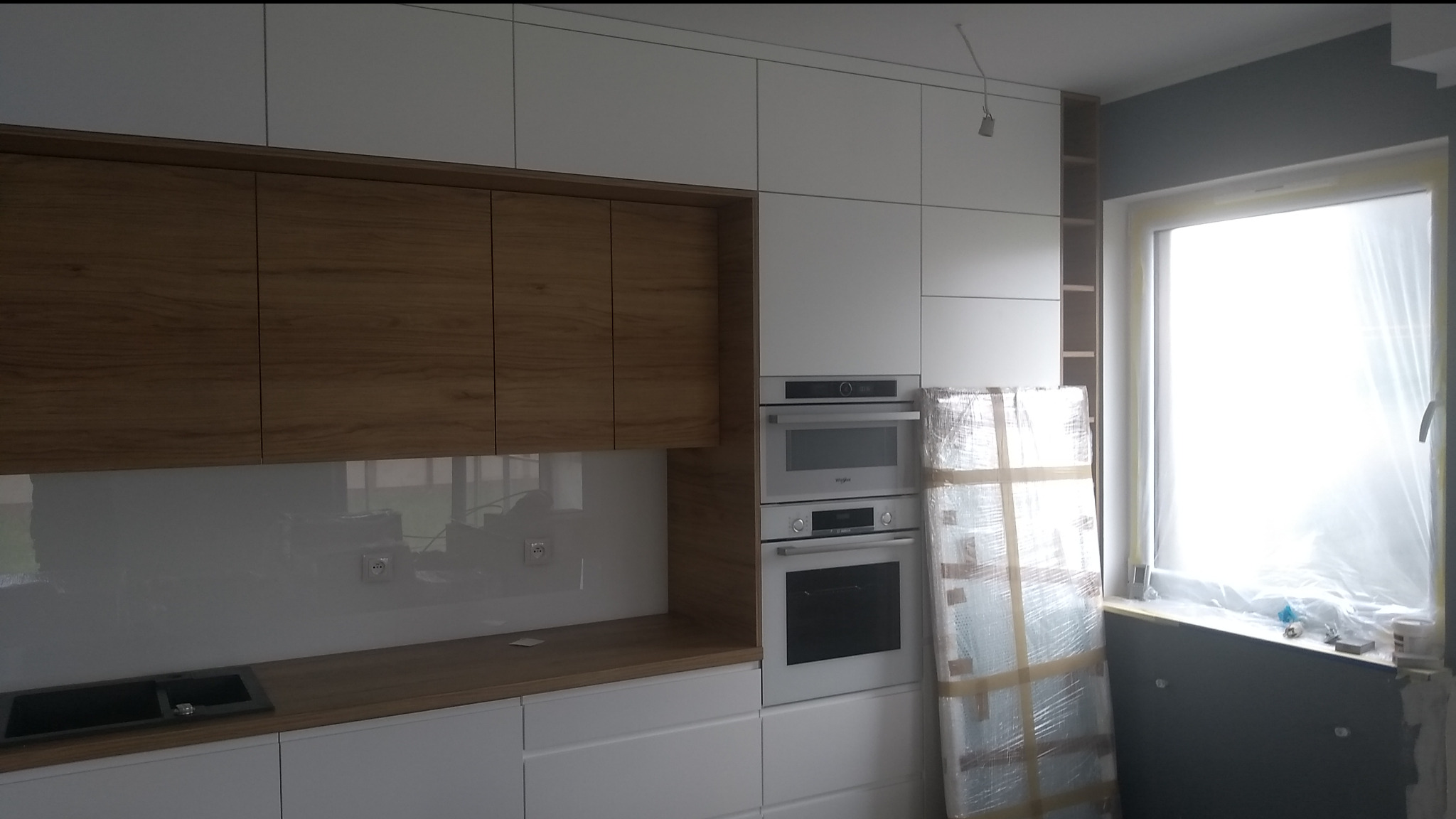 Nowoczesna kuchnia z białymi frontami i drewnianymi szafkami wiszącymi. Widoczne sprzęty AGD w zabudowie i okno zabezpieczone folią malarską.