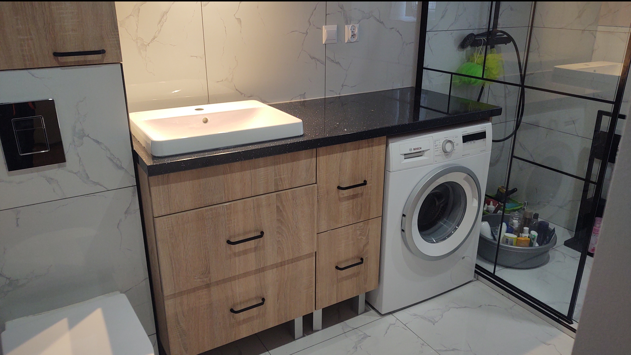 Nowoczesna łazienka z szafką pod umywalkę z frontami w kolorze drewna, czarnym blatem i pralką Bosch. Kabina prysznicowa z czarnymi profilami.