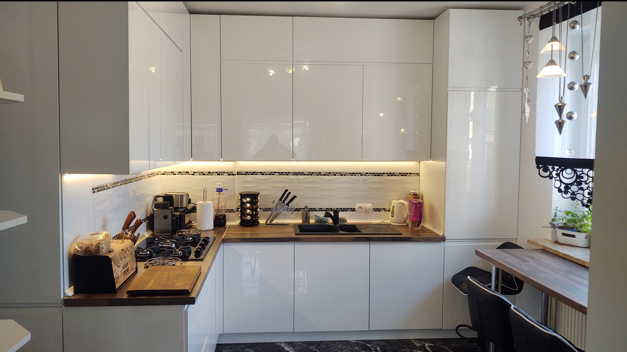 Biała, nowoczesna kuchnia z połyskiem, oświetleniem LED pod szafkami i drewnianym blatem. Widoczny zlew, płyta grzewcza i dekoracyjne zasłony w oknie.