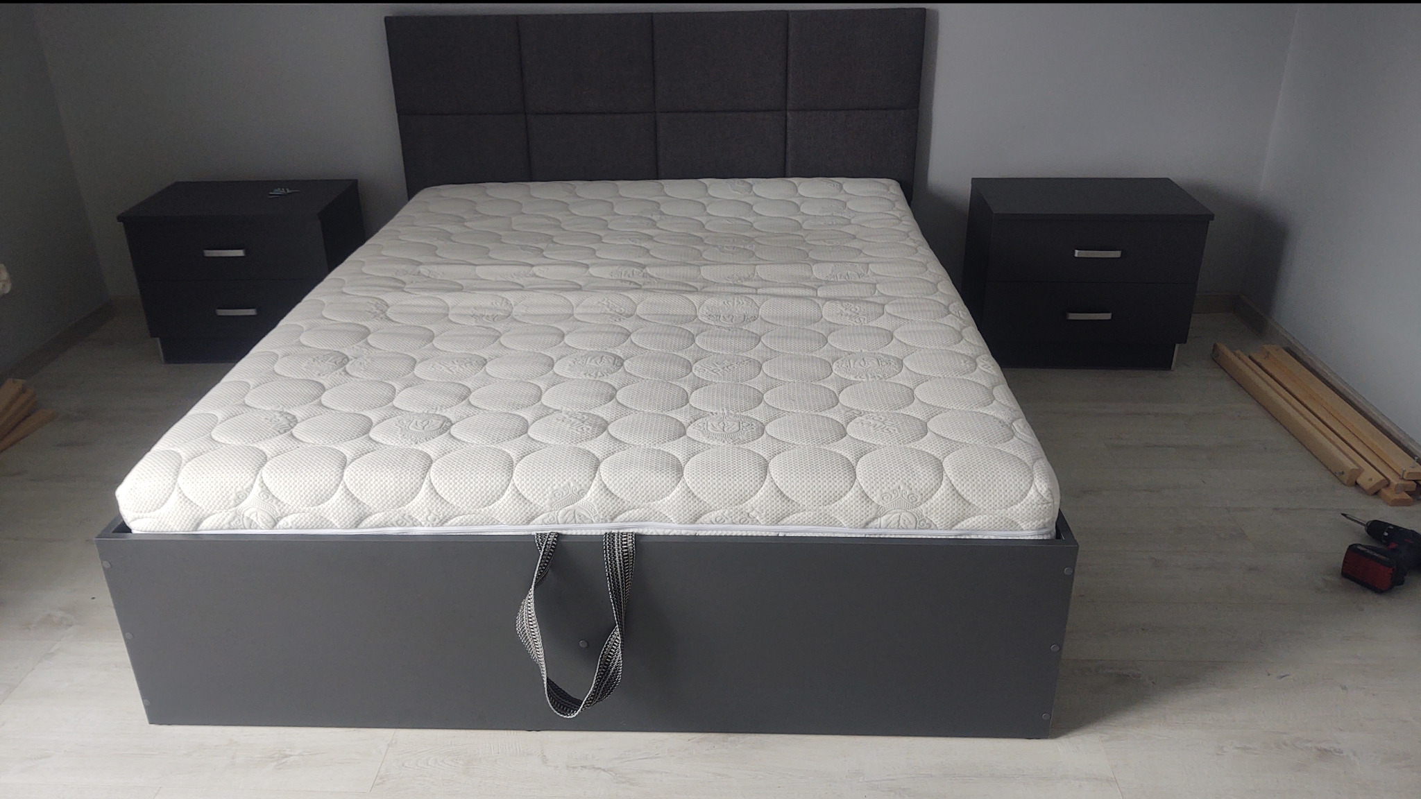 Szare łóżko z tapicerowanym zagłówkiem i materacem, z szafkami nocnymi po bokach. W tle ściana i podłoga w odcieniach szarości. Widoczne narzędzia i elementy montażowe.