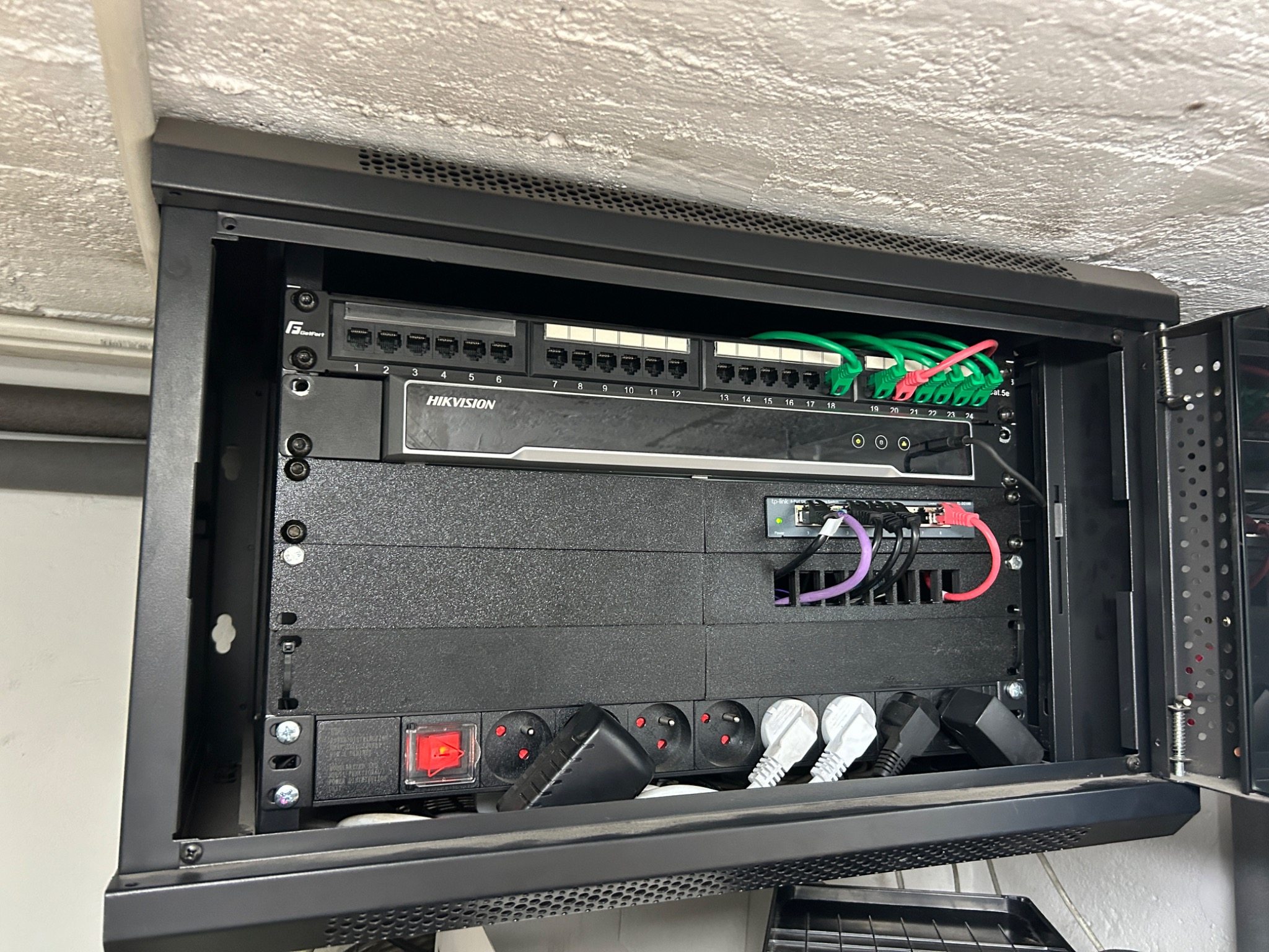 Zamknięta szafa rack z patch panelem, switchem i listwą zasilającą Hikvision. Widoczne kable sieciowe w kolorach zielonym, fioletowym i czerwonym.