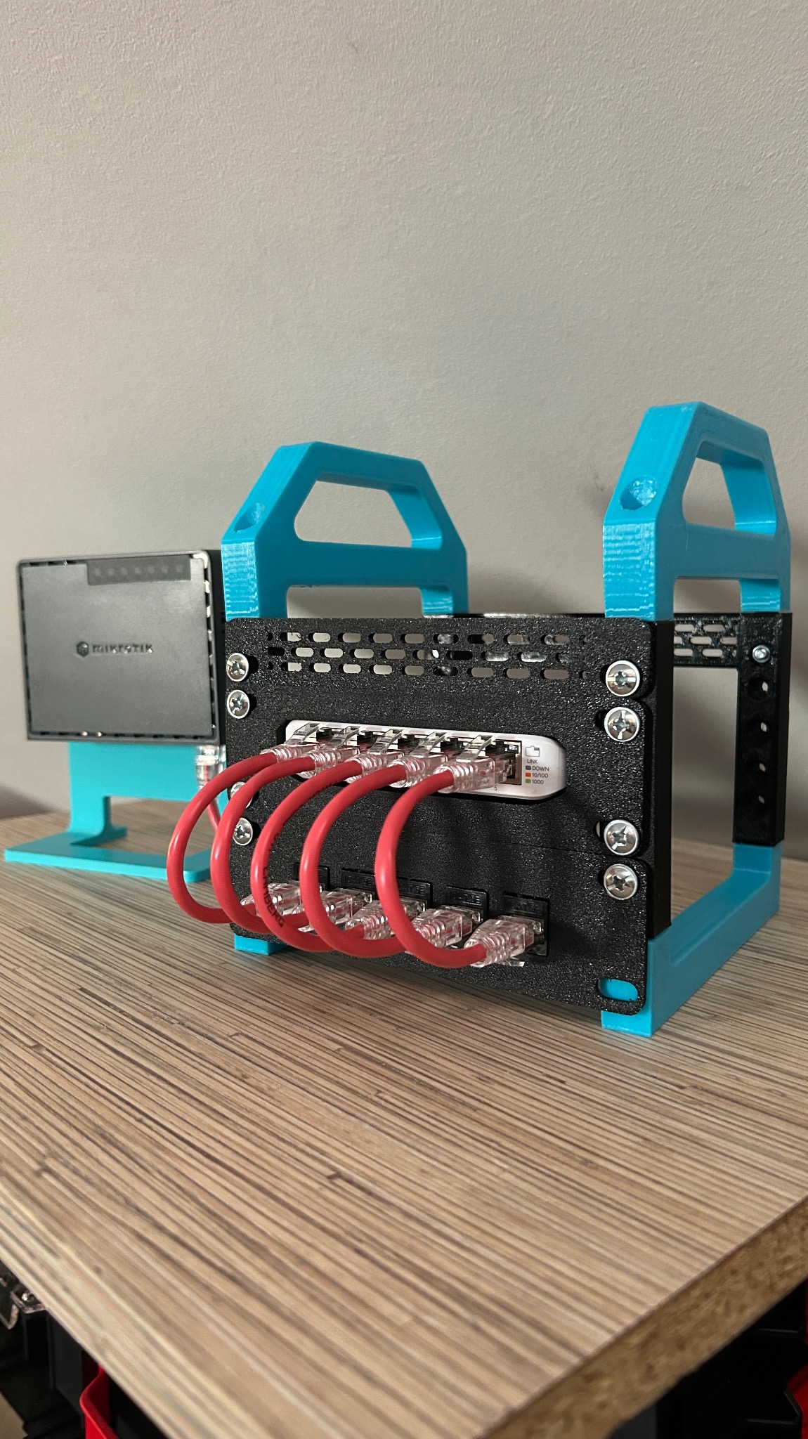 Switch sieciowy Mikrotik w obudowie 3D z turkusowymi uchwytami i czerwonymi kablami Ethernet, na drewnianym blacie. Widoczne diody LED i śruby mocujące.