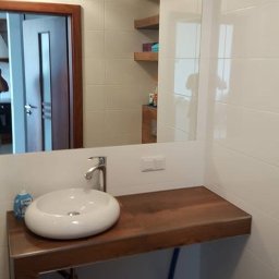 Finish Expert Daniel Brzozowski - Nowoczesna łazienka: białe płytki, drewniany blat z umywalką, duże lustro z oświetleniem. Widoczne półki w odbiciu lustra i brązowa podłoga.