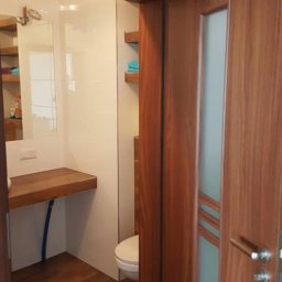 Finish Expert Daniel Brzozowski - Nowoczesna łazienka z drewnianymi akcentami: podłoga, blat umywalki i drzwi z przeszkleniem. Białe płytki i minimalistyczne półki dopełniają wystrój.