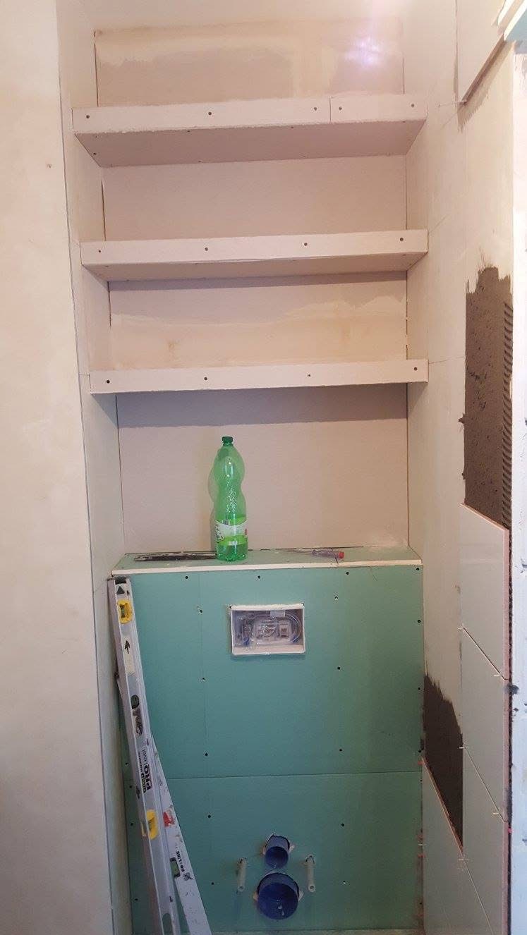 Niewykończona łazienka: zabudowa z płyt gipsowo-kartonowych, przygotowane półki i częściowo położone płytki. Widoczny poziomica i butelka z wodą.