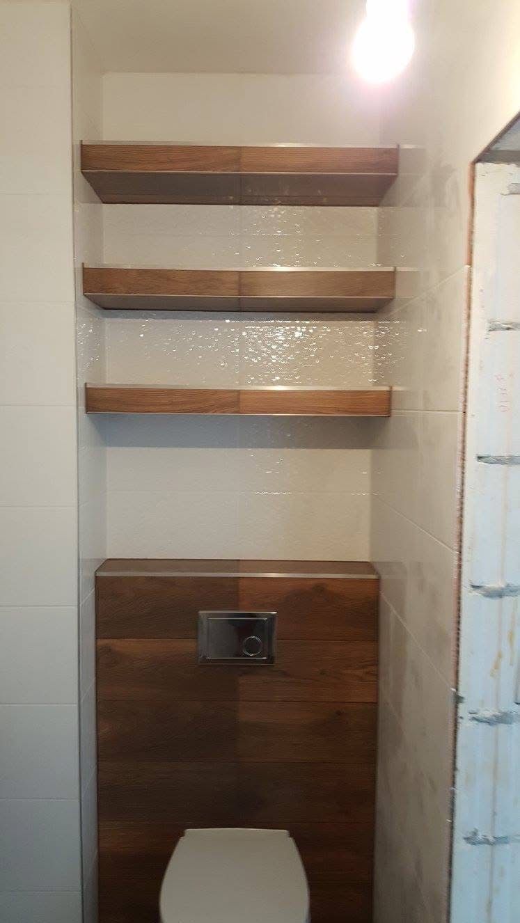 Wykończona łazienka z podwieszaną toaletą, obudową z paneli drewnopodobnych i trzema półkami z tego samego materiału. Białe płytki na ścianach, nowoczesny styl.