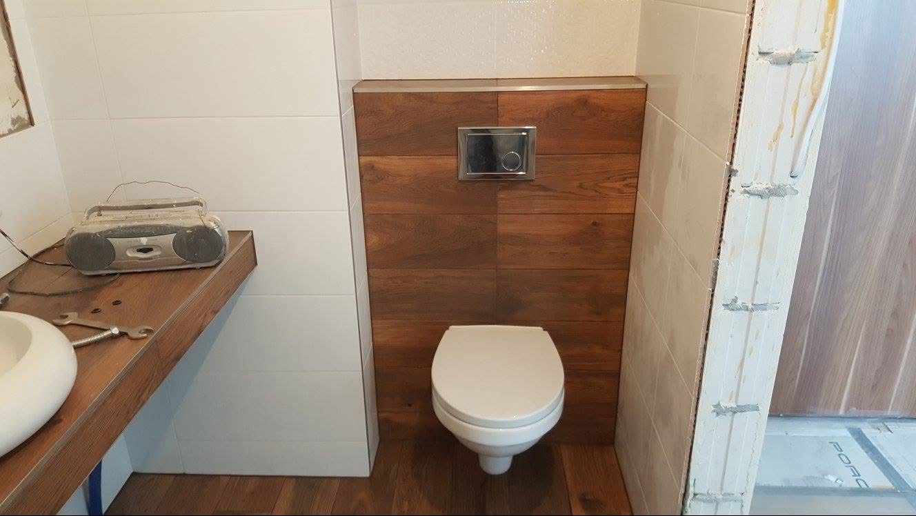 Nowoczesna toaleta podwieszana z drewnianą obudową i chromowanym przyciskiem spłukującym, białe płytki na ścianach, drewniany blat z radiem retro.