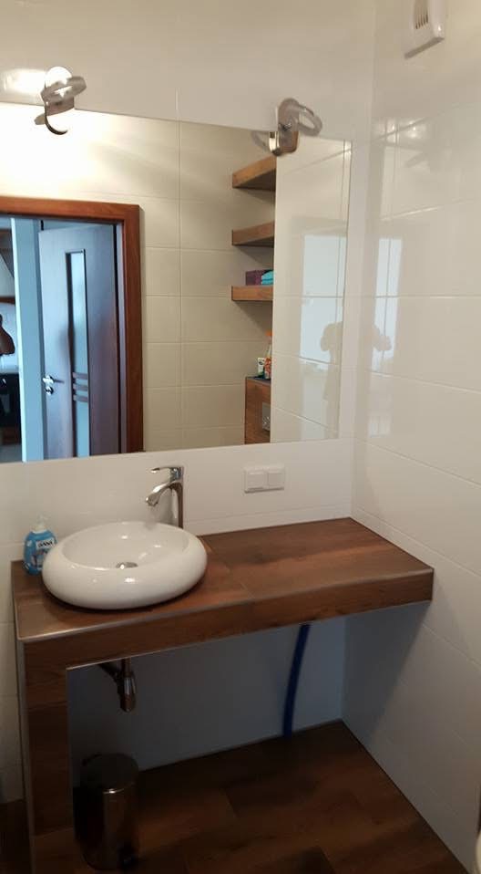 Nowoczesna łazienka: białe płytki, drewniany blat z umywalką, duże lustro z oświetleniem. Widoczne półki w odbiciu lustra i brązowa podłoga.