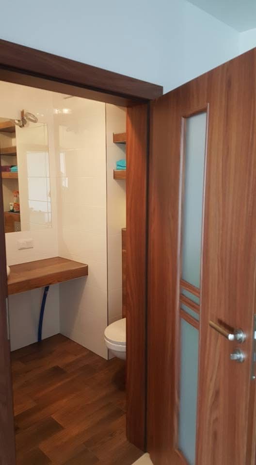 Nowoczesna łazienka z drewnianymi akcentami: podłoga, blat umywalki i drzwi z przeszkleniem. Białe płytki i minimalistyczne półki dopełniają wystrój.