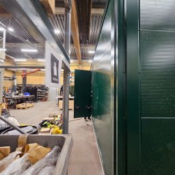 Madbud Magdalena Lisiak - Wnętrze hali produkcyjnej z widoczną ścianą z zielonych płyt warstwowych i otwartymi drzwiami. Widoczne elementy konstrukcyjne i wyposażenie hali.