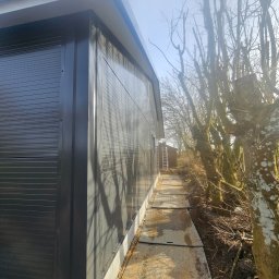 Madbud Magdalena Lisiak - Nowoczesna hala z płyt warstwowych w kolorze grafitowym, widok z boku. Na pierwszym planie ścieżka z metalowych płyt, w tle drzewa bez liści i drabina oparta o budynek.