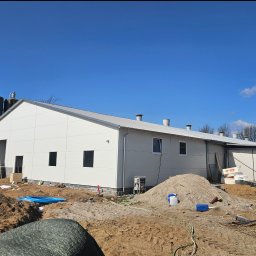 Madbud Magdalena Lisiak - Nowoczesna hala z białych płyt warstwowych w trakcie budowy, widoczne silosy w tle, ziemia i materiały budowlane na pierwszym planie, błękitne niebo.