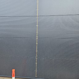 Madbud Magdalena Lisiak - Szara ściana hali z płyt warstwowych, widoczne łączenia i mocowania. Na dole leżą deski, a obok stoi pomarańczowy pachołek. Detal montażu.