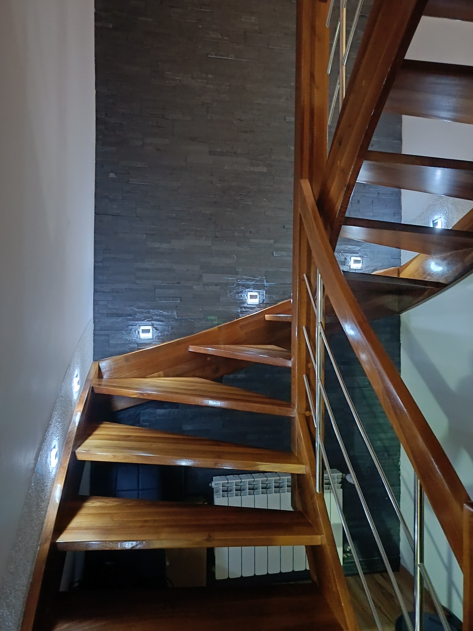 Drewniane schody spiralne z metalową balustradą, oświetlone punktowo, na tle ściany wykończonej kamieniem dekoracyjnym w odcieniach szarości. Widoczny grzejnik.