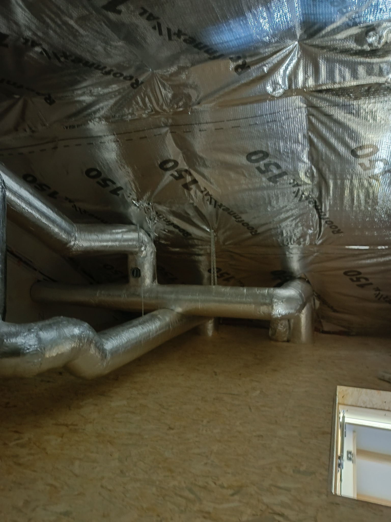 Izolacja poddasza z folią aluminiową i rurami wentylacyjnymi. Widok na drewnianą konstrukcję i okno dachowe. Detal wykończenia izolacji.