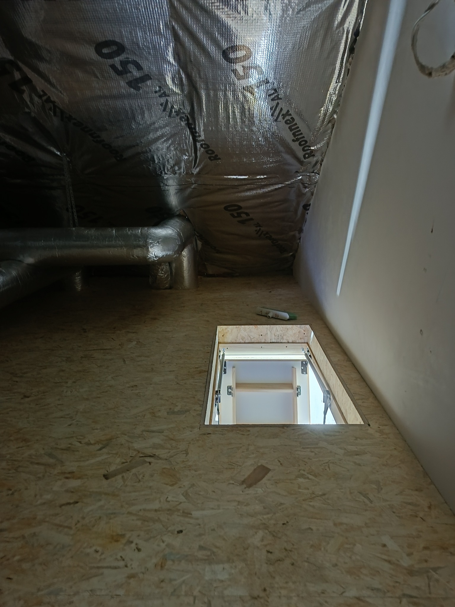 Ocieplone poddasze z widoczną izolacją termiczną, fragmentem instalacji wentylacyjnej i otworem do włazu. Wykończona podłoga z płyty OSB.