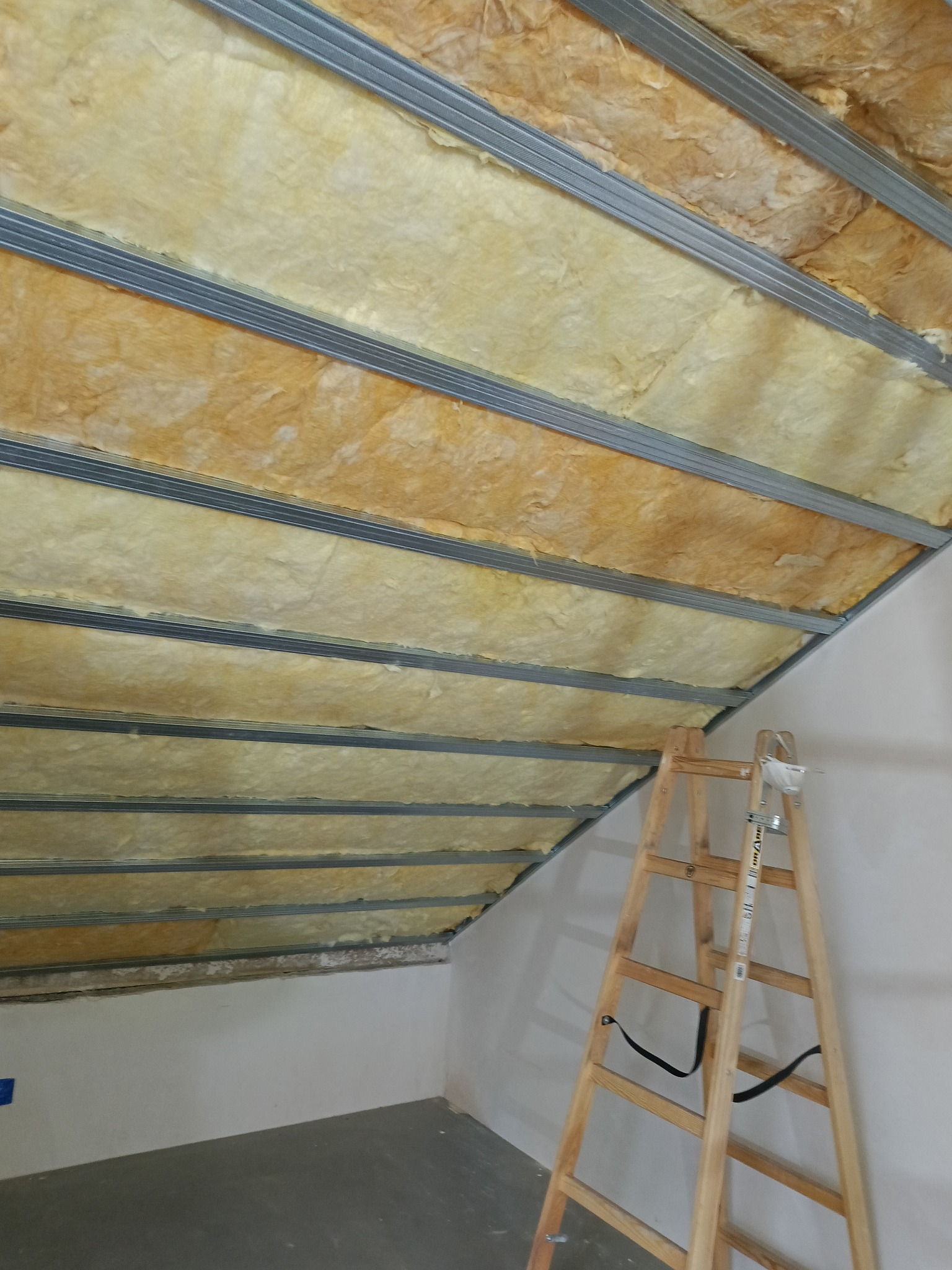 Ocieplenie poddasza wełną mineralną, widoczne metalowe profile i drabina. Izolacja termiczna w trakcie montażu, sufit skośny.