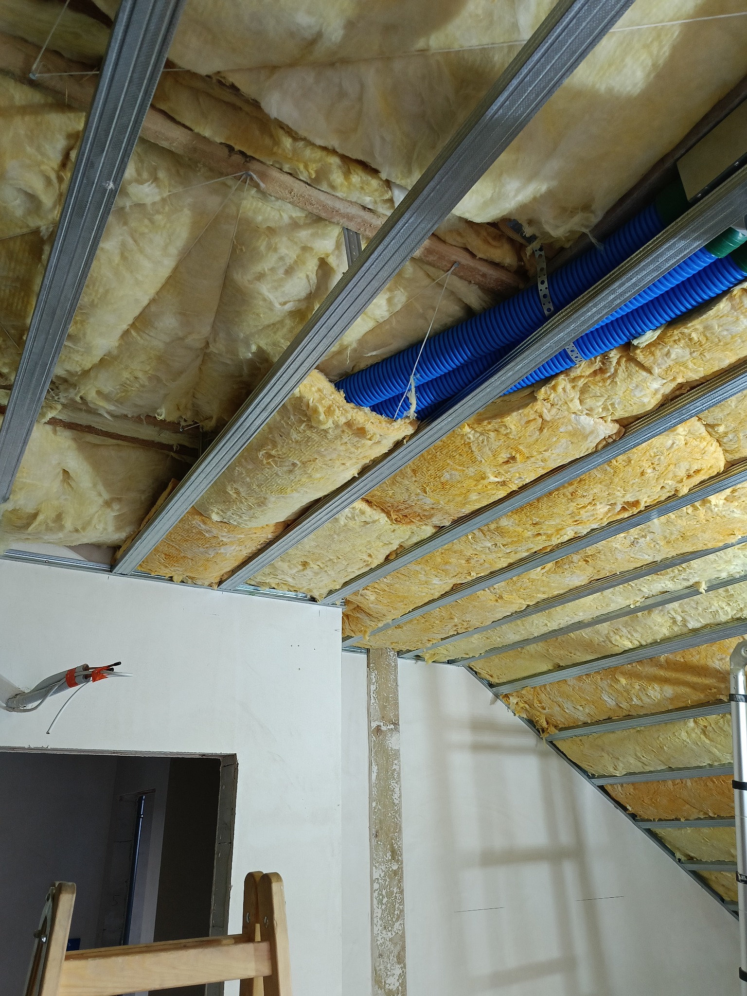 Ocieplenie poddasza wełną mineralną, widoczne profile metalowe i niebieskie rury instalacyjne. Częściowo zamontowana płyta gipsowa. Prace w toku, widok z dołu.