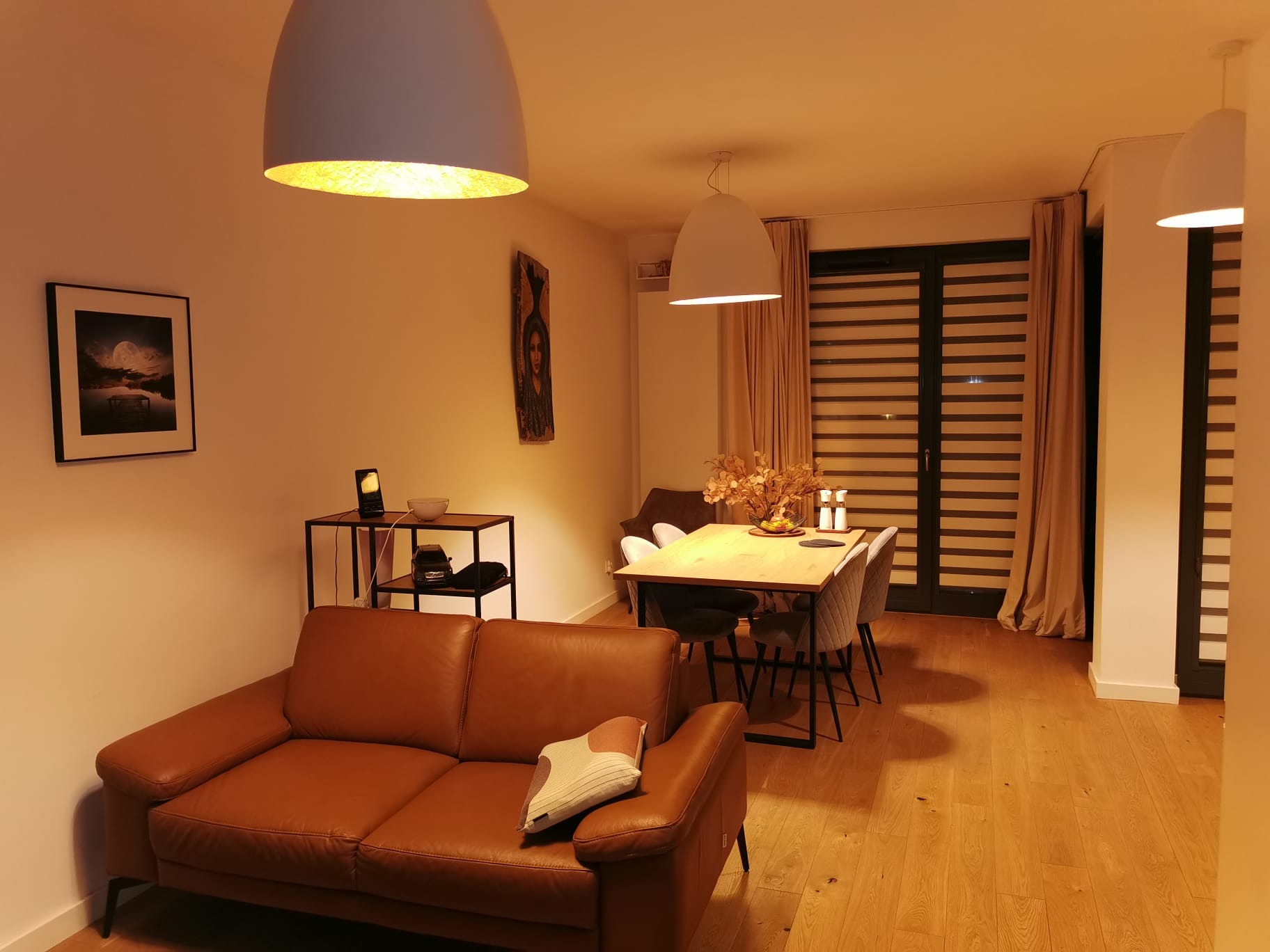 Ciepłe wnętrze salonu z brązową skórzaną sofą, drewnianym stołem jadalnym i trzema lampami wiszącymi. Na ścianie obrazy i dekoracje. Parkiet na podłodze.