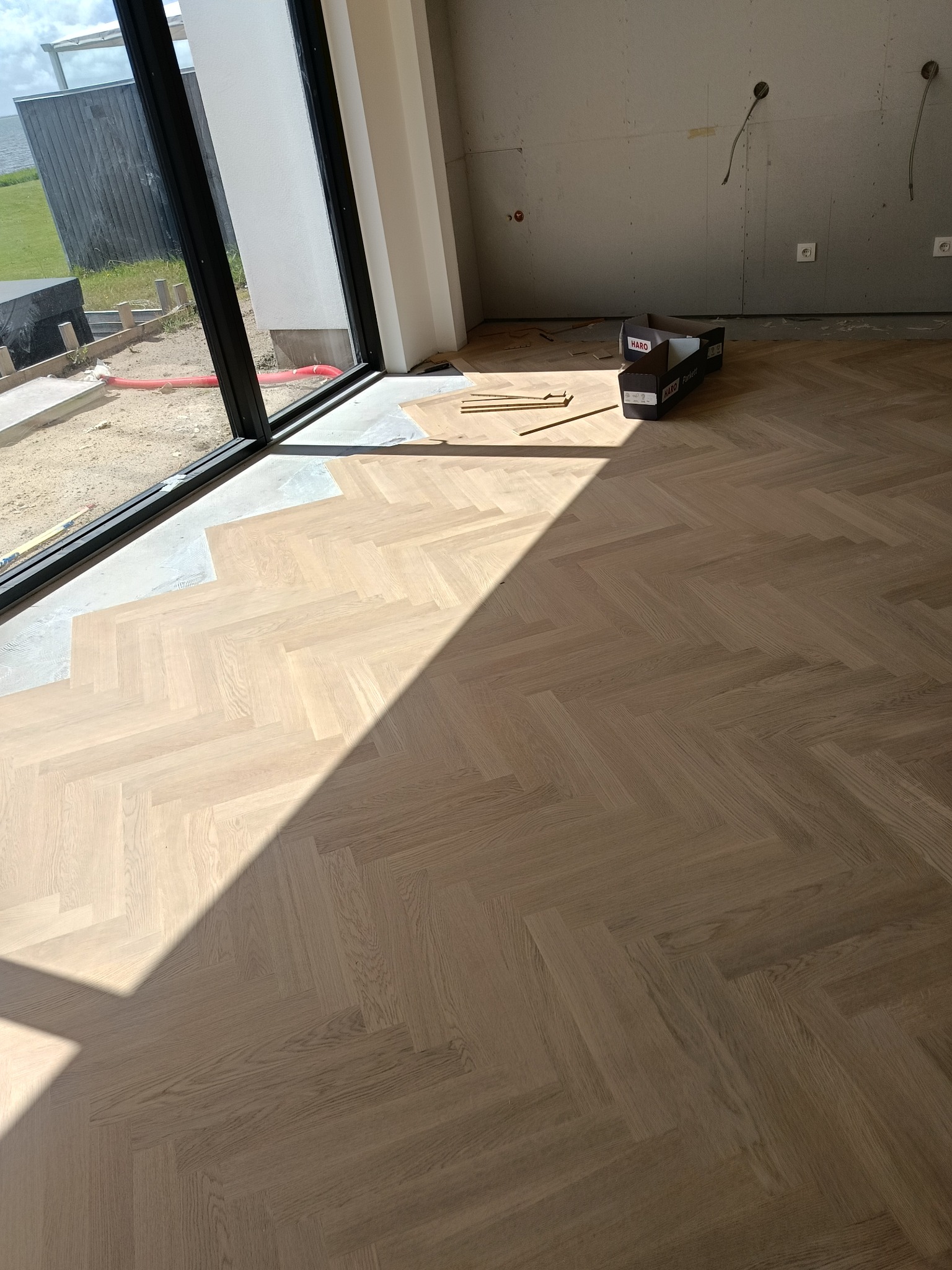 Jasny parkiet ułożony we wzór jodełki francuskiej w nowym wnętrzu z dużym oknem. Częściowo położona podłoga, widoczne kartony parkietu i przygotowane podłoże.