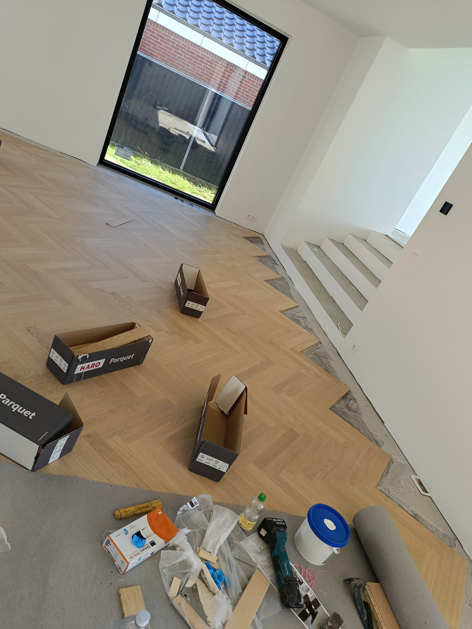 Układanie parkietu w jodełkę francuską, widoczne narzędzia i materiały montażowe, częściowo ułożona podłoga, okno w tle. Jasne wnętrze w trakcie wykańczania.