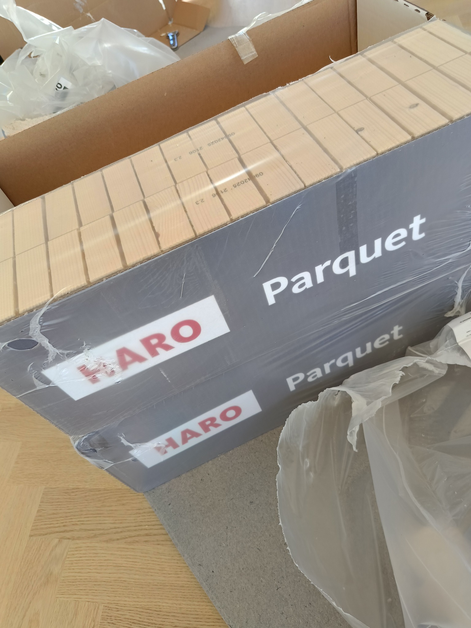 Paczka parkietu Haro, widoczna struktura drewna, opakowanie z folii. Podłoże z ułożonych paneli. Kartonowe pudło w tle.