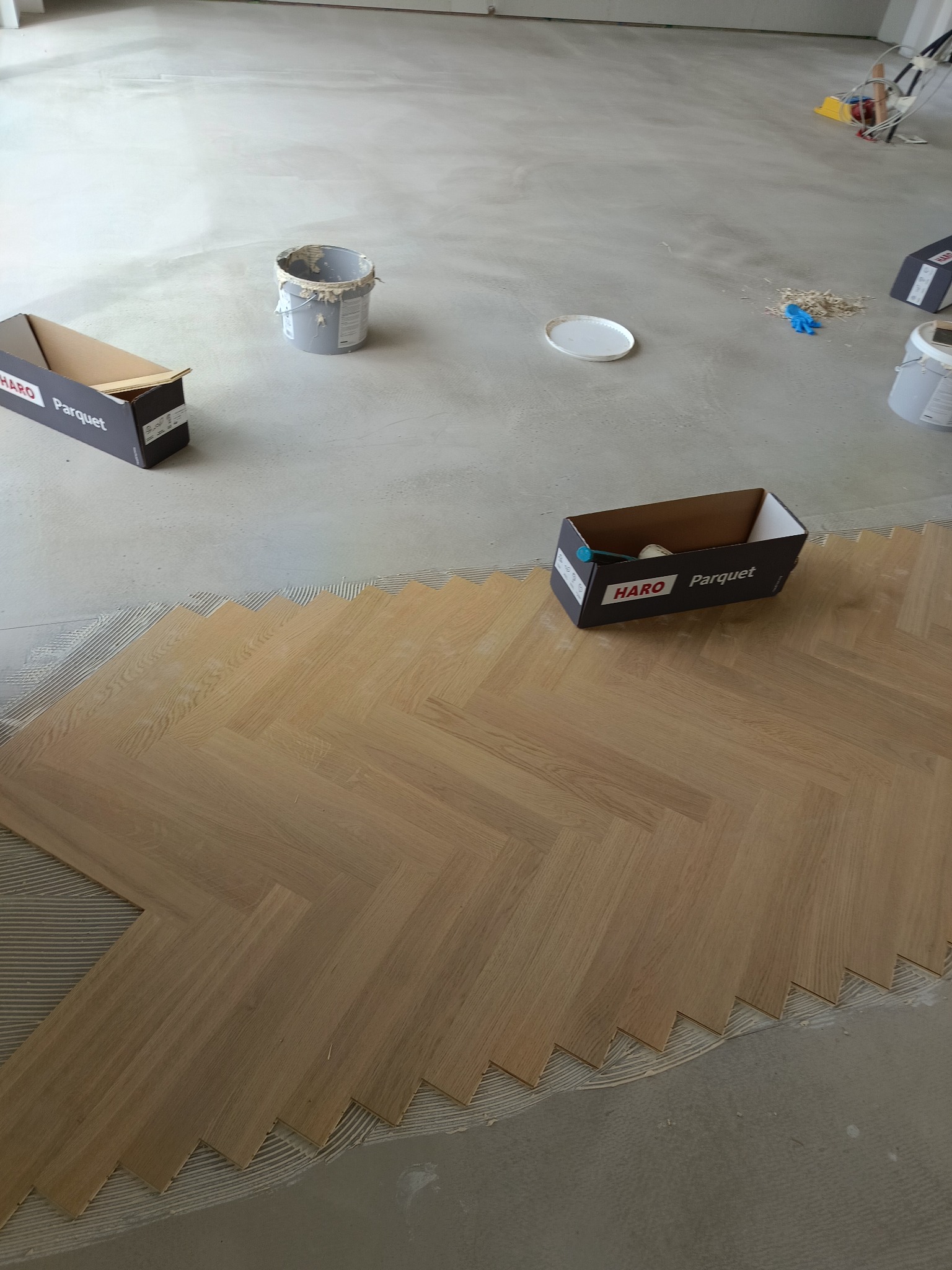 Układanie parkietu w jodełkę: fragment podłogi z ułożonym parkietem, widoczne narzędzia i materiały budowlane, w tle surowa posadzka.