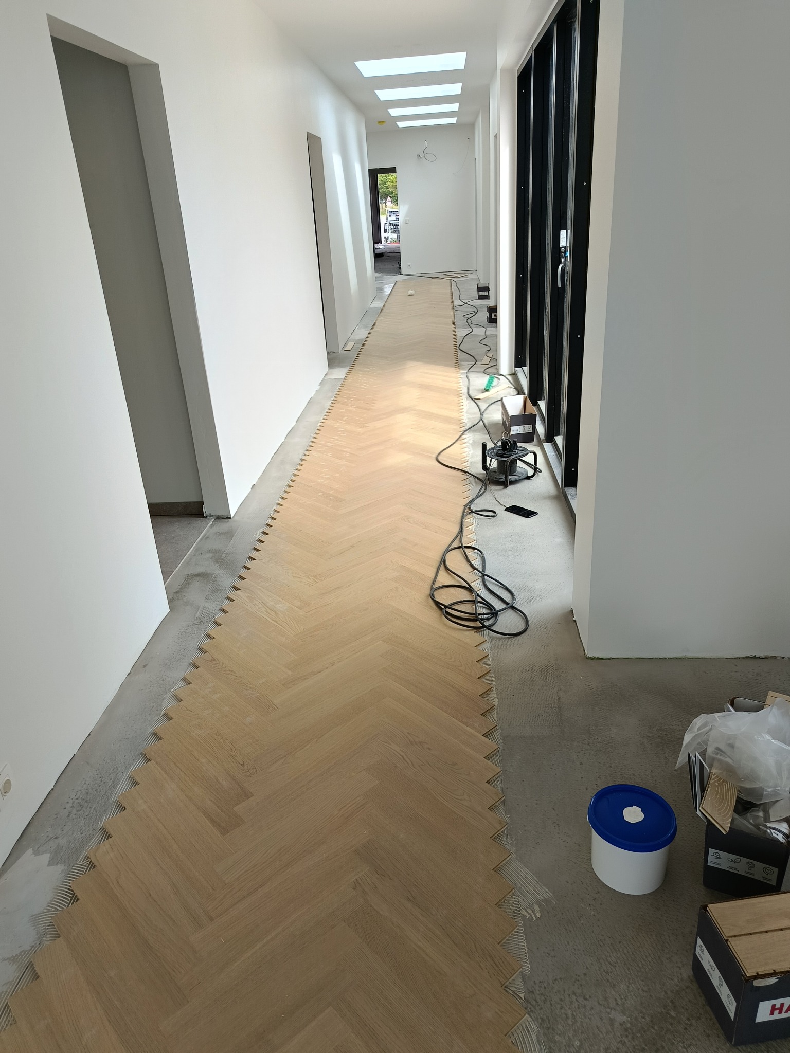 Układanie parkietu w jodełkę w długim korytarzu z białymi ścianami i oknami. Widoczne narzędzia i materiały budowlane. Częściowo ułożona podłoga z jasnego drewna.