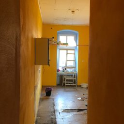 Paintpol - Żółte ściany w trakcie remontu, widoczna drabina, okno z niebieską ramą i fragment podłogi z usuniętymi panelami. W tle szafka na ścianie i wiadro.