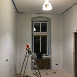 Paintpol - Pokój po szpachlowaniu, przygotowany do malowania. Widoczne zabezpieczenia taśmą malarską, drabina, grzejnik, surowe ściany i podłoga. Lampa sufitowa oświetla wnętrze.