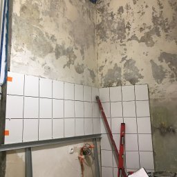 Paintpol - Układanie białych płytek w rogu pomieszczenia z surowymi ścianami. Widoczne poziomice, listwa montażowa i notatki. Prace glazurnicze w trakcie realizacji.
