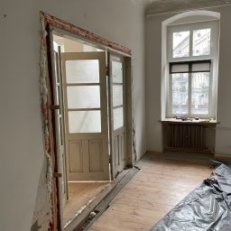 Paintpol - Demontaż ościeżnicy w jasnym pokoju z drewnianą podłogą, zabezpieczoną folią. Widoczne ślady pianki montażowej i odsłonięta konstrukcja ściany. Okno z widokiem na ulicę.