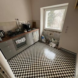 Paintpol - Kuchnia w trakcie prac wykończeniowych: czarno-biała podłoga w karo, widoczne narzędzia i materiały budowlane przy oknie. Częściowo wyposażona kuchnia.