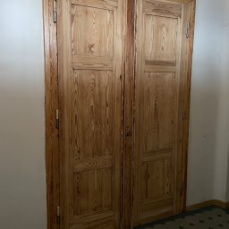 Paintpol - Drewniane, dwuskrzydłowe drzwi wewnętrzne z widocznym usłojeniem, zamontowane w narożniku pomieszczenia z ciemną podłogą w geometryczny wzór.