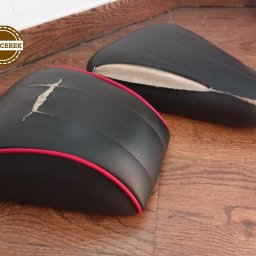 Dwa czarne siedziska, jedno z czerwoną lamówką i pęknięciem, drugie z widocznym wypełnieniem, leżą na drewnianej podłodze. Widoczne ślady zużycia.