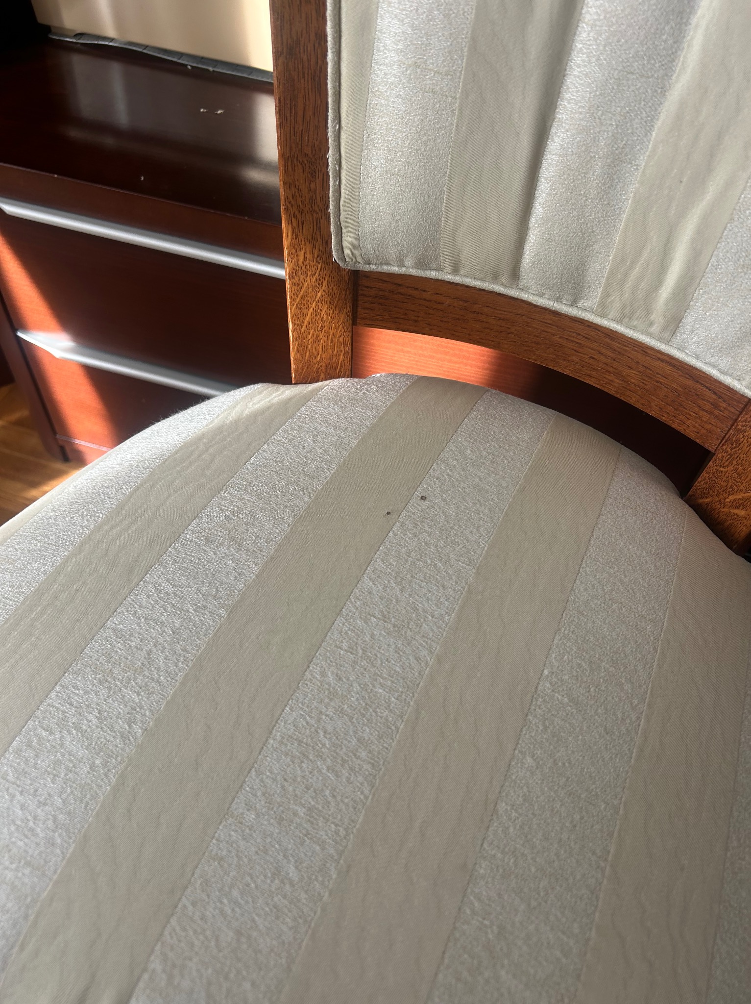 Krzesło z tapicerką w beżowe pasy, widoczne drobne zabrudzenia na siedzisku. Drewniana rama krzesła w ciepłym odcieniu brązu. W tle fragment komody.