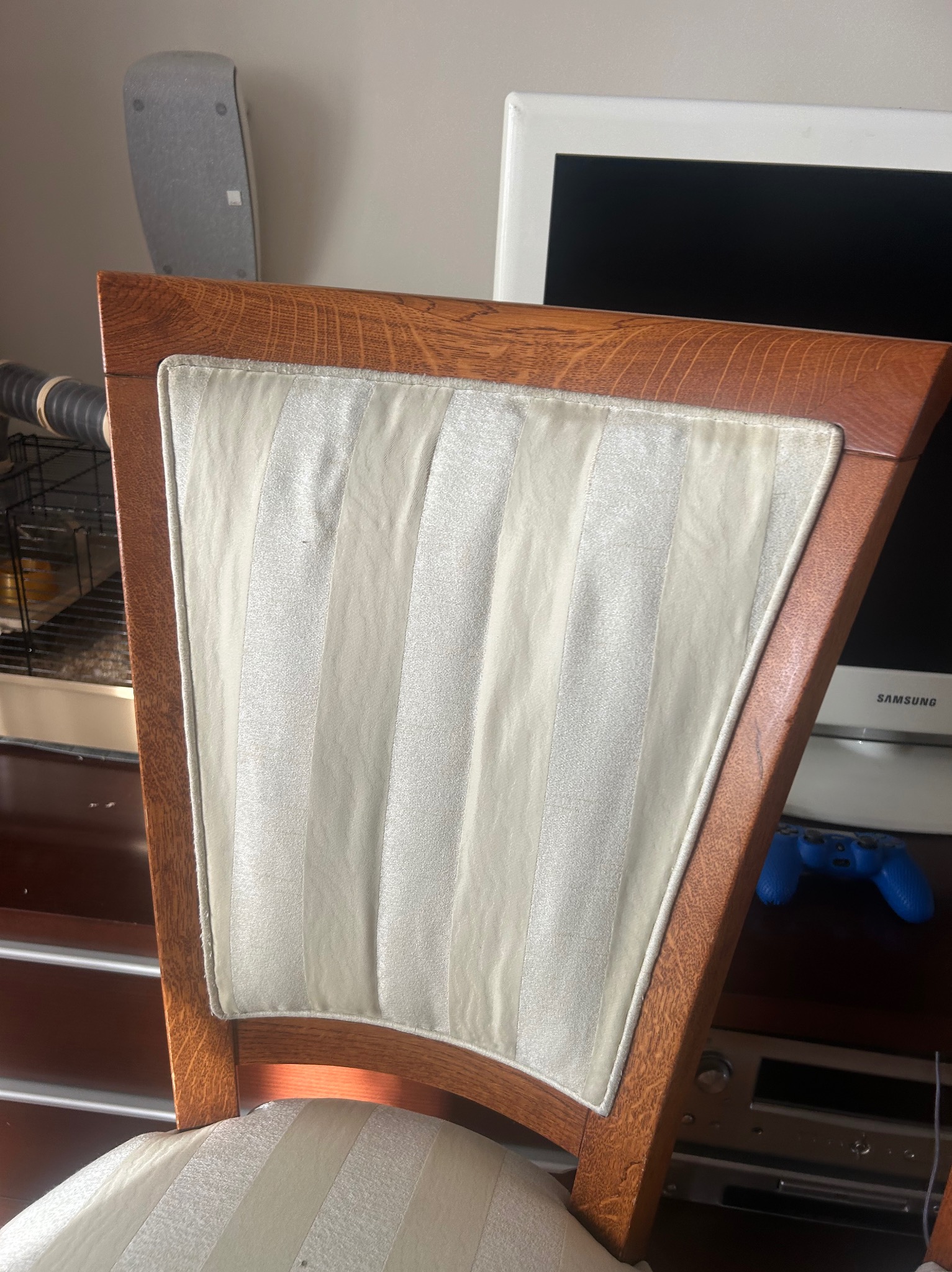 Krzesło z drewnianą ramą i tapicerowanym oparciem w paski, widziane od tyłu, na tle sprzętu RTV i klatki dla zwierząt. Widoczne detale tapicerki i struktura drewna.