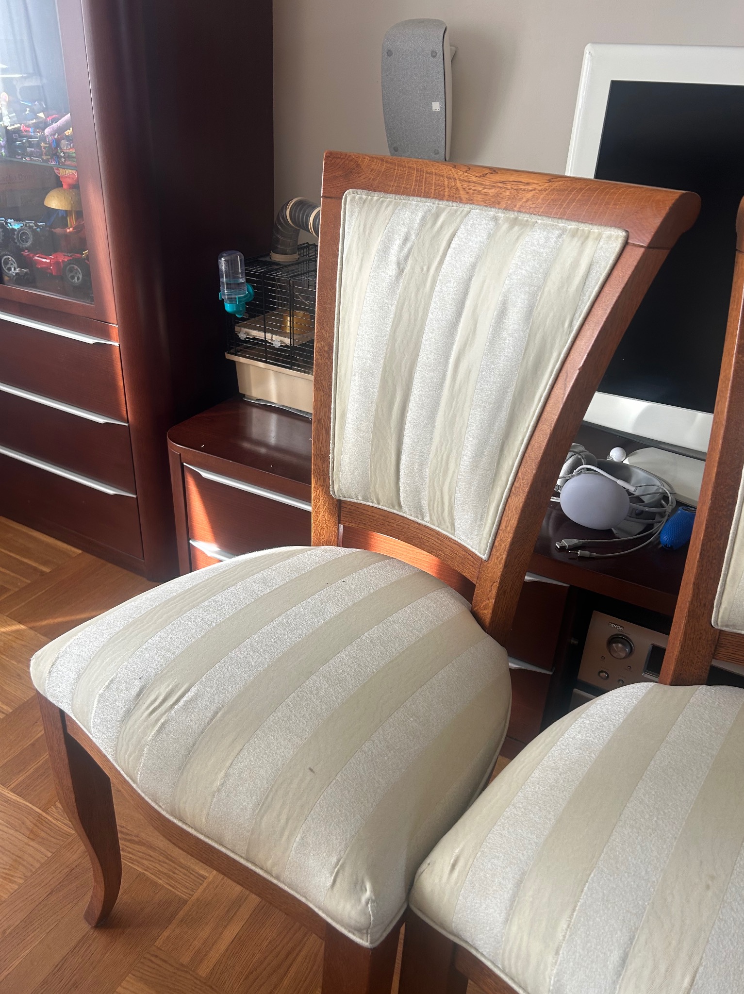 Dwa krzesła z drewnianą ramą i tapicerką w kremowe pasy, ustawione w pokoju z parkietem. Widoczny fragment szafki z zabawkami i klatka dla zwierząt w tle.