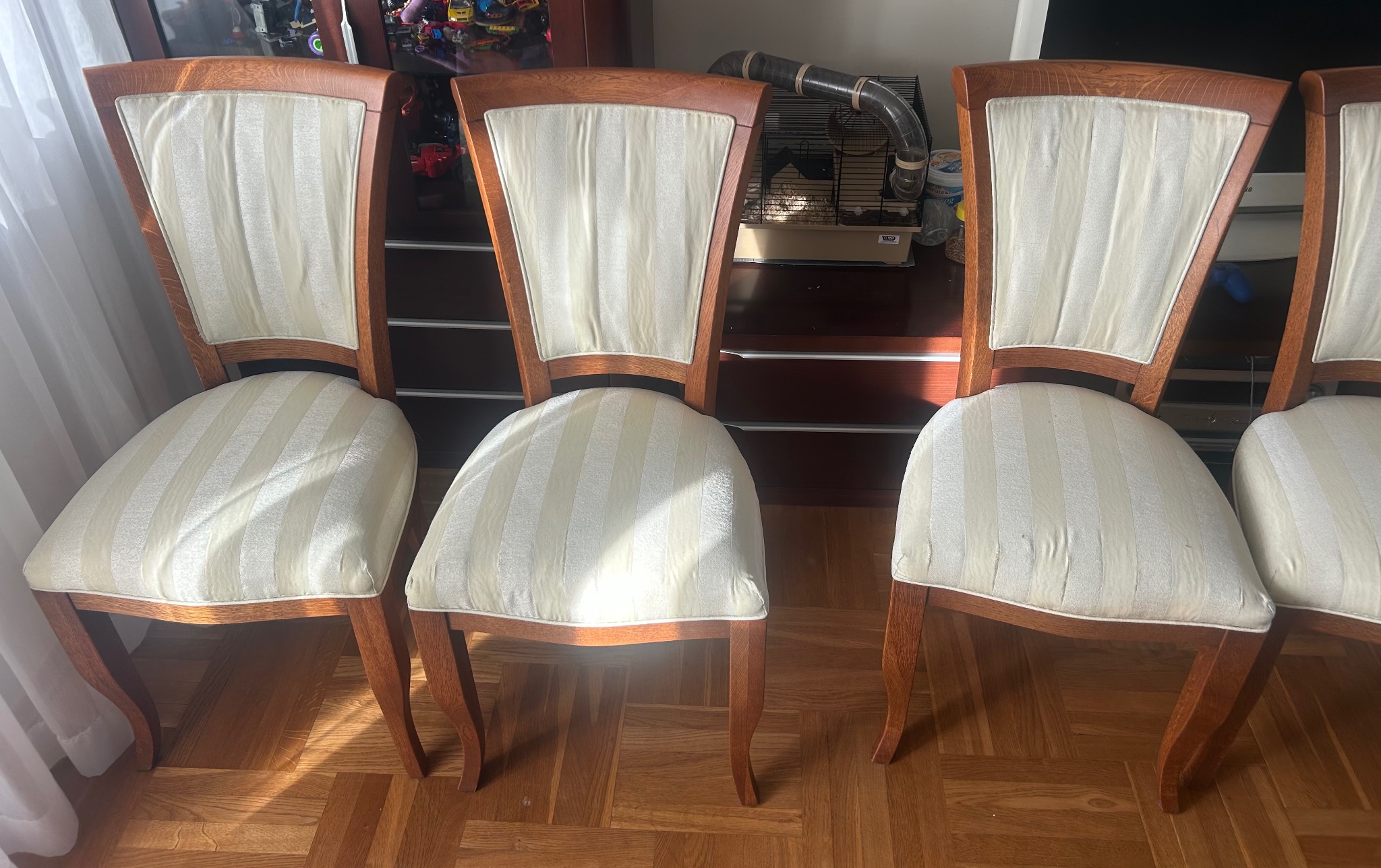 Cztery krzesła z drewnianą ramą i tapicerką w kremowe pasy, ustawione na parkiecie. W tle komoda i klatka dla gryzonia z tunelem.