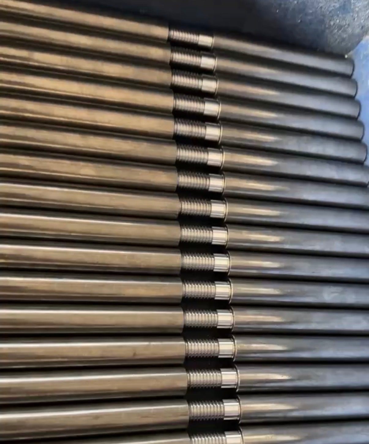 Rząd precyzyjnie wykonanych, metalowych prętów z gwintowanymi końcówkami, ułożonych równolegle. Widoczne detale obróbki i metaliczny połysk.