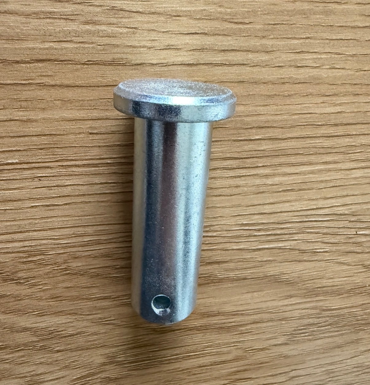 Metalowy trzpień z otworem, leżący na drewnianej powierzchni. Detal wykonany z precyzją, widoczne ślady obróbki.