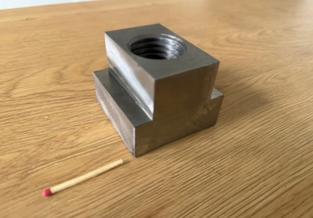 Metalowy element z gwintem, obrobiony z precyzją, leżący na drewnianej powierzchni obok zapałki dla skali. Widoczne ślady obróbki.