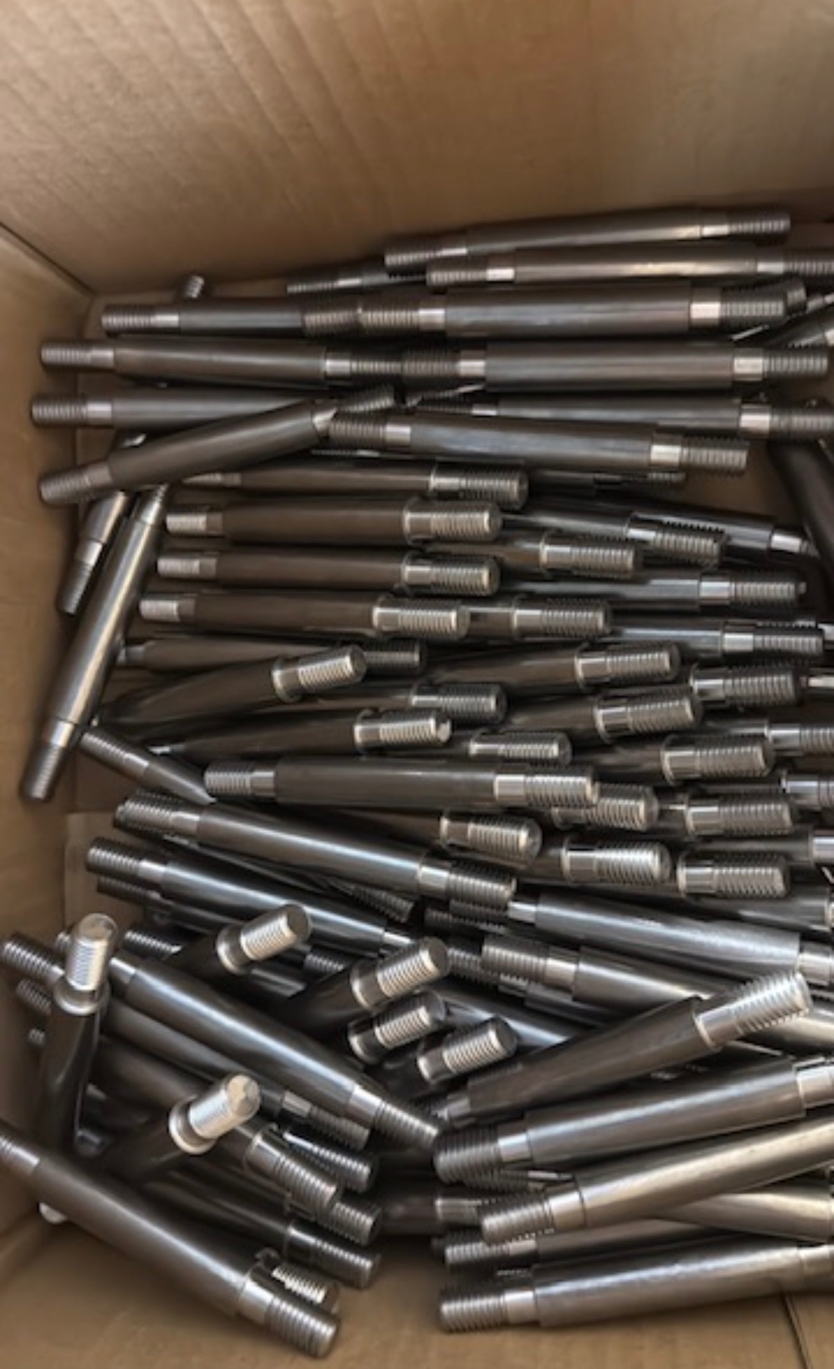 Partia metalowych, obrobionych wałków z gwintem na końcach, ułożonych w kartonowym pudle. Widoczne ślady obróbki.